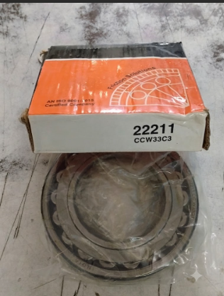 A large metal bearing, sealed in clear plastic, is placed in front of its orange and white paper packaging box, which features printed text including 22211 CCW33C3.