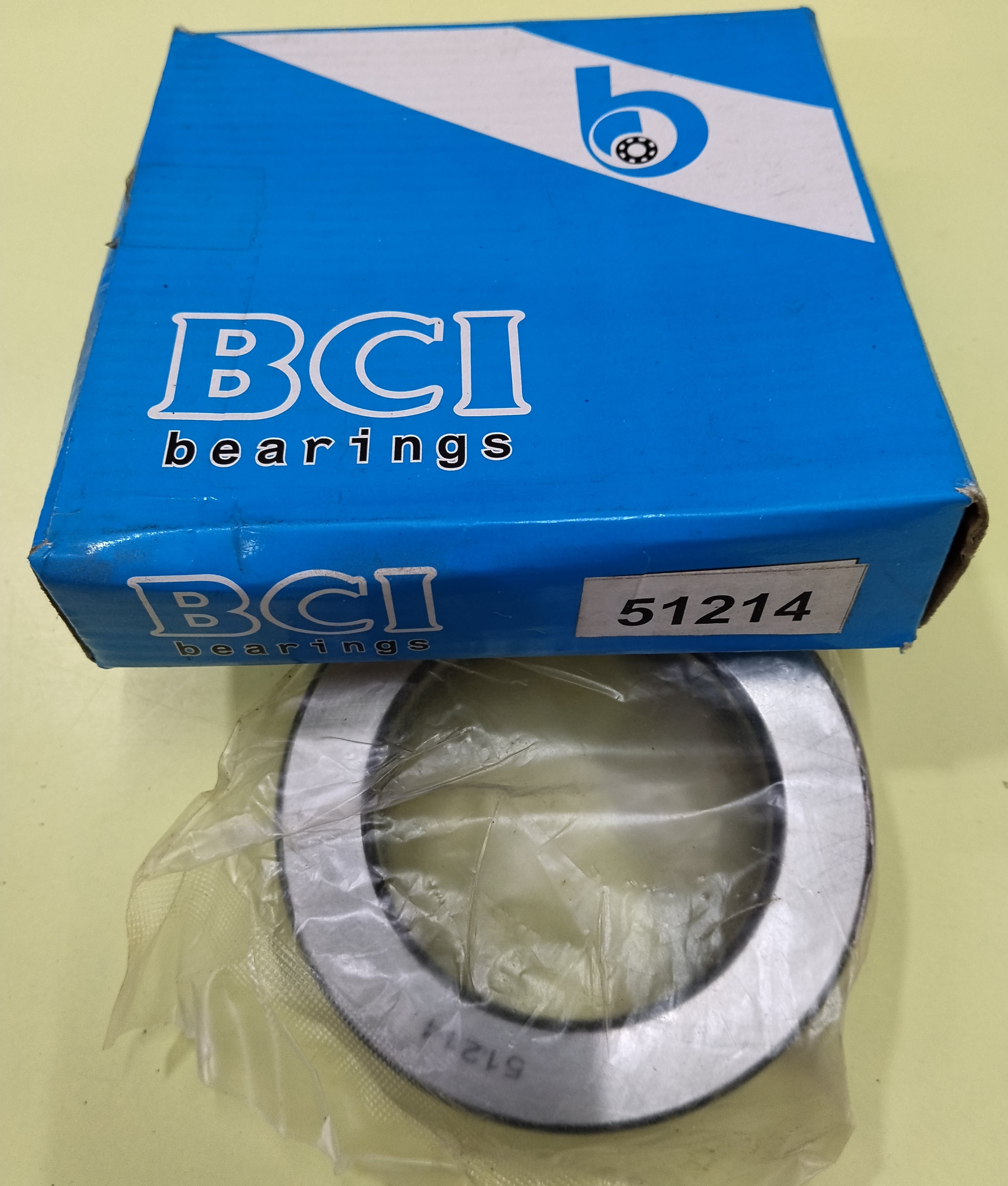 A BCI bearings ring-shaped 51214 THRUST BALL bearing in clear plastic packaging, positioned in front of its blue and white BCI Bearings product box on a pale yellow surface.