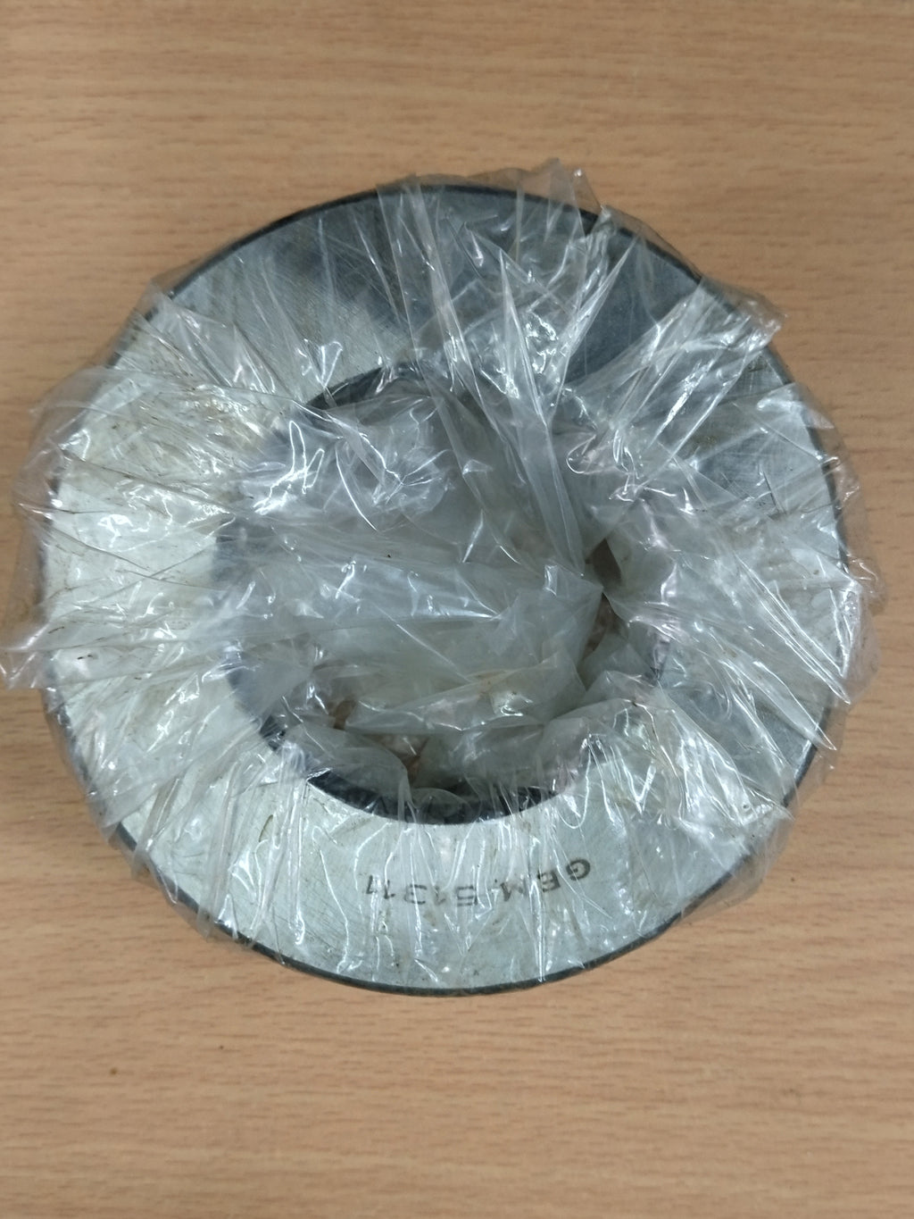 A circular GEM 51311 metal bearing wrapped in clear plastic, placed on a light wood surface.