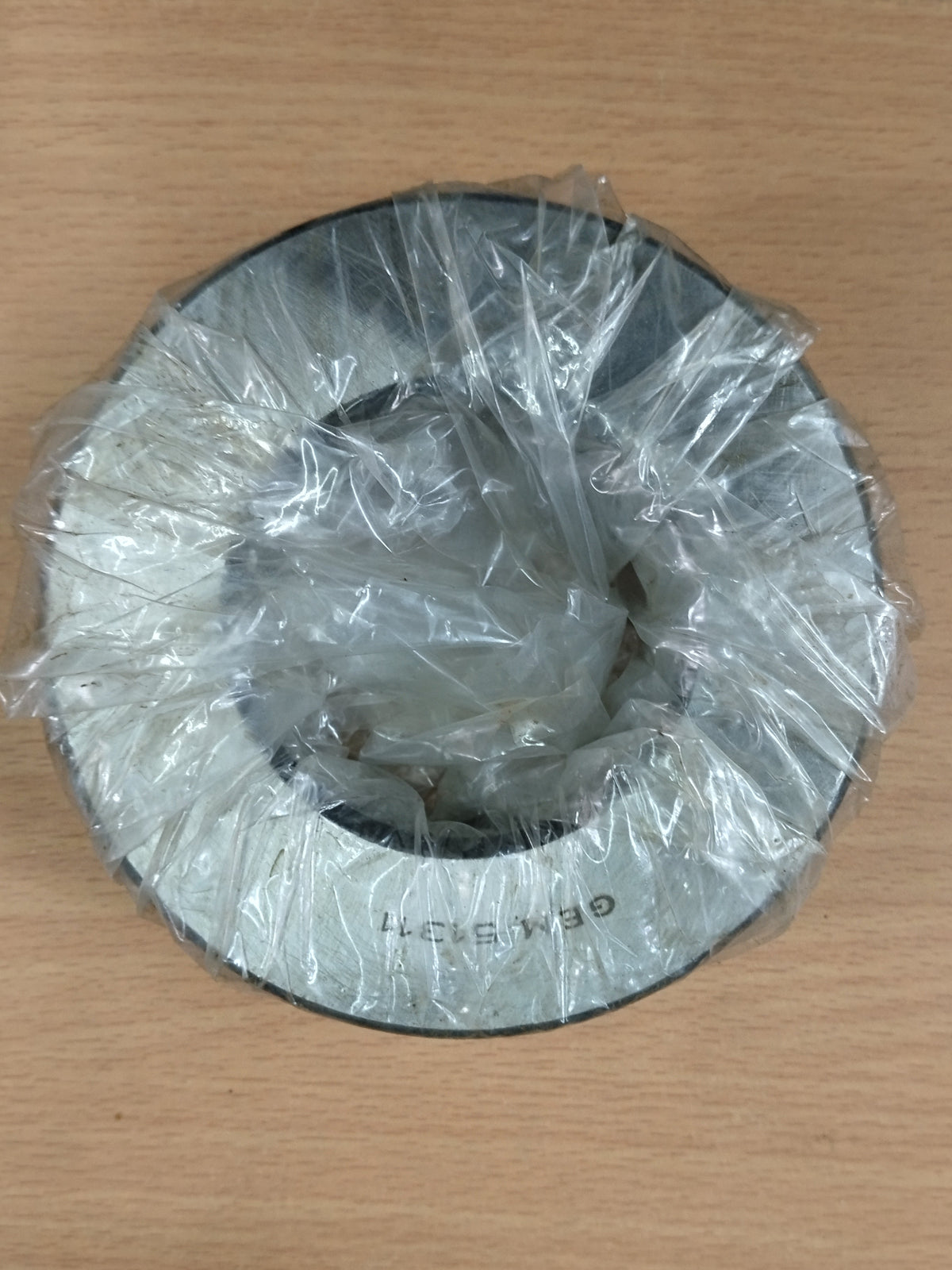 A circular GEM 51311 metal bearing wrapped in clear plastic, placed on a light wood surface.