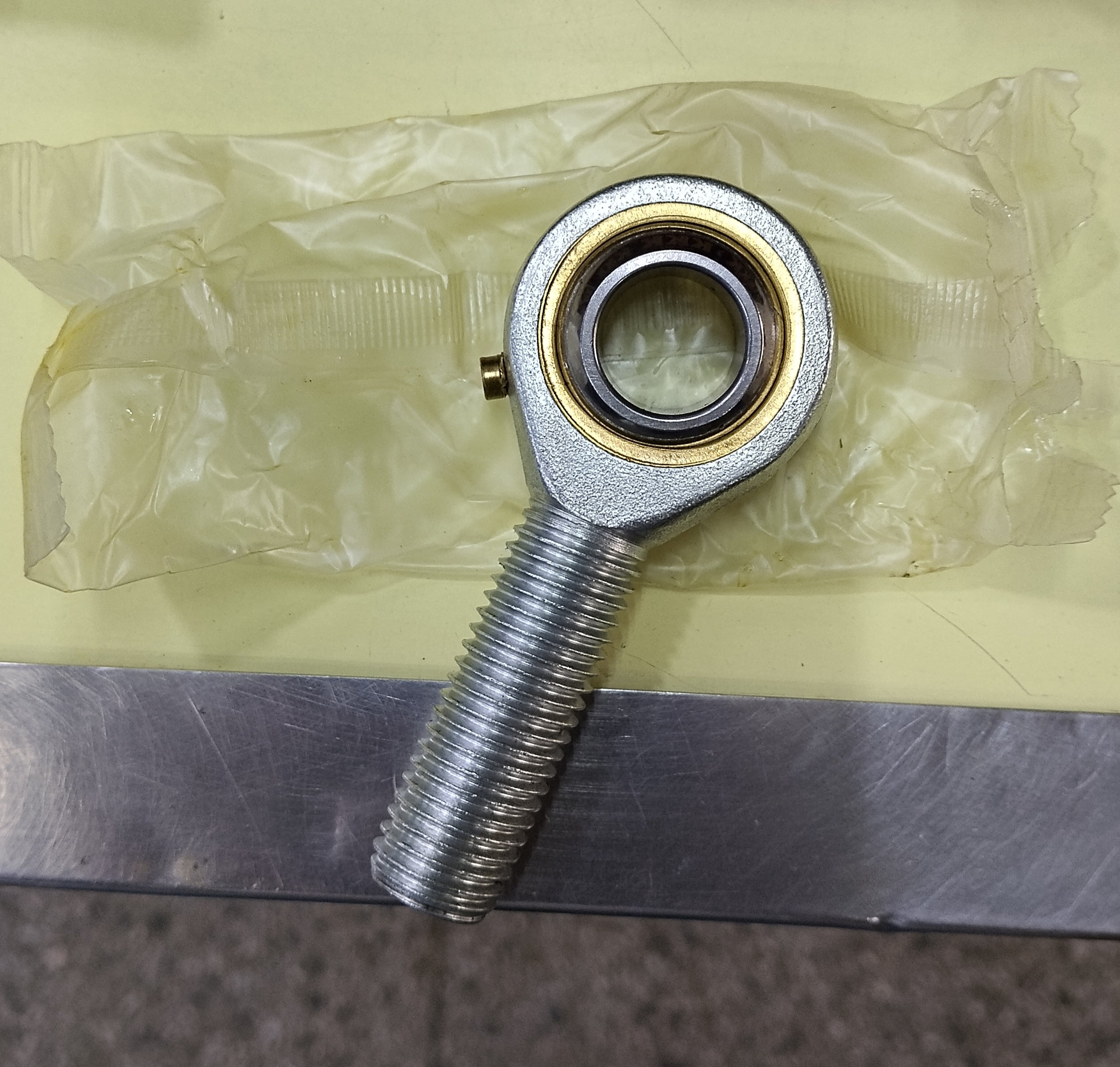A metal threaded rod joint bearing with a silver ball head and gold-toned inner ring, resting on a translucent plastic bag over a yellow surface. POS 16 POS rod end bearings